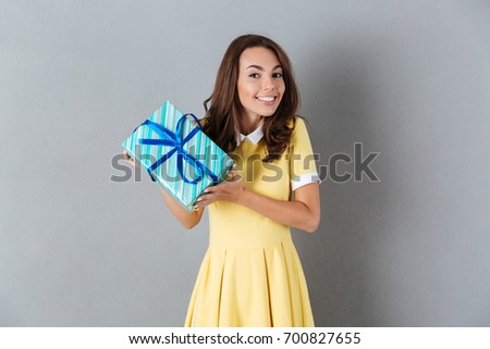 Picture of emotional smiling young caucasian lady standing over grey background holding gift. Looking camera.
