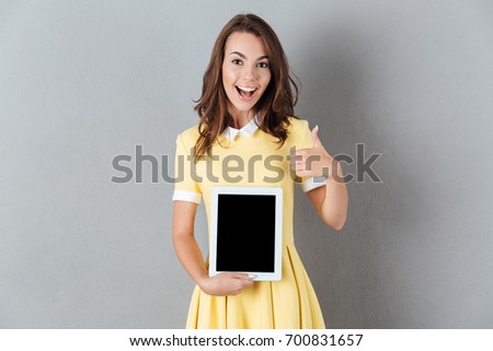Image of cheerful young caucasian lady showing display of tablet computer. Looking camera showing thumbs up.