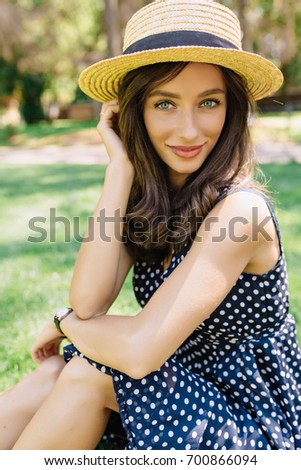 Photo of style woman wearing summer hat and cute dress in the summer park. She is sitting on the green grass and smiles. Great emotions. Summer day. Background green park.