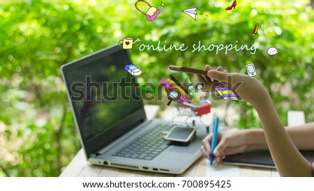 On line shopping concept. Hands holding smart phone with doodle icons shopping while writing order on notepad and recording data in laptop computer on wood table in home garden green background