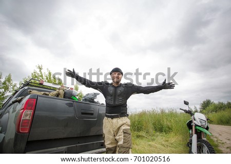 Happy smiling young man with arms wide open outdoors wearing motorcycle gear and looking at the camera, truck with helmets and dirt bike on the scenic background.