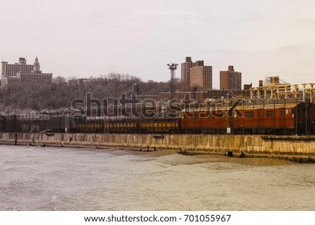 Urban landscape of NYC (USA), a red and grey suburb train rolling alongside the river. Buildings in the background