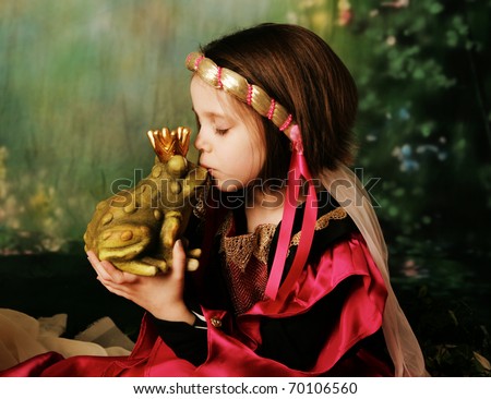 Portrait of a cute young preschool girl dressed as a princess in a pink and gold gown, posing and kissing a frog prince wearing a crown