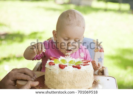 One year old girl sitting near birthday cake and eating it on the garden.