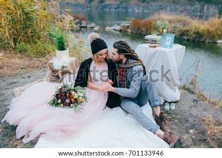 Stylish couple newlyweds are relaxing on a plaid and sitting before a lake. Bride and groom with dreadlocks are posing on nature. Autumn wedding ceremony outdoors. Full length portrait