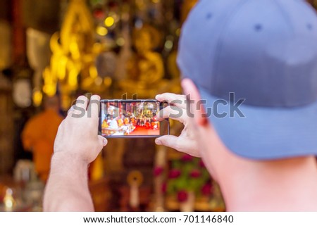 The caucasian man wearing blue cap is taking picture by cell phone to the Buddhist monks while they are chanting in the golden Buddha images hall in the temple.