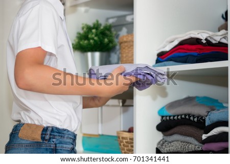 The European blond boy teenager brings wardrobe order, puts everything in its place, hides things in boxes. Wardrobe with women's, men's and child's clothing. Closet. Royalty-Free Stock Photo #701341441
