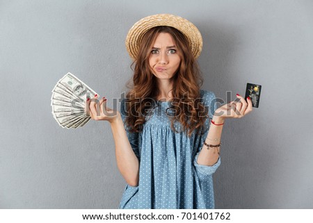 Picture of confused young woman standing over grey wall wearing hat holding money and credit card.