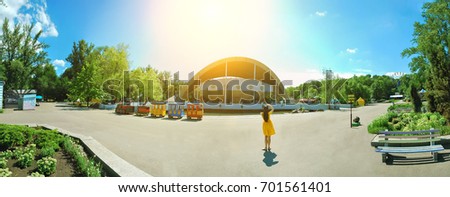 Panorama of the city park with a summer theater and a lake.A tall girl in a yellow dress photographs a panorama of a tall old theater building on the lake shore.Hot, sunny summer