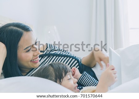 A little cute girl lying with her mother on the white bed in the bedroom, daughter hug her teddy bear and watching cartoon by tablet with her mom, rest time, happy family concept 