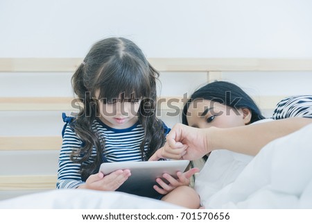 A little cute girl lying with her mother on the white bed in the bedroom, daughter watching cartoon by tablet with her mom, rest time, happy family concept 