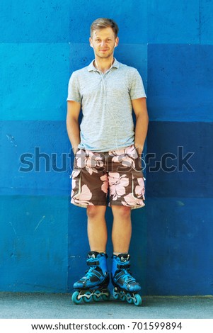 Portrait of happy handsome young roller in inline skates standing against blue wall.