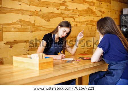 Portrait of two pensive young women sitting at a wooden table. Girls baristas in an apron are waiting for customers in a cafe. Coffee business concept. Small local business.