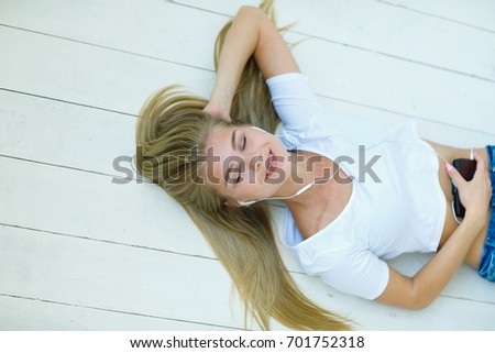 Portrait of a resting beautiful young girl with blond hair in a white T-shirt and blue jeans, lying on a wooden floor and listening to her favorite music on her smartphone. lifestyle concept. 