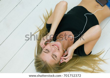 Portrait of a young and happy girl with blond hair in a black T-shirt listening to her favorite song through headphones while lying on the wooden floor . People, youth and lifestyle concept. 