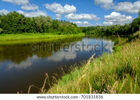 Bank of Gauja River in Latvia with blue sky and white clouds reflecting in water