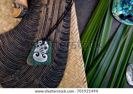New Zealand - Maori themed objects - carved wooden mere and metal and greenstone pendant with flax leaves and abalone - paua shells on gray stone background
