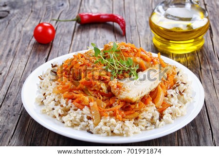 portion of delicious white fish braised with carrot, onion, tomato sauce, spices and herbs with wild and long grain rice on white plate on dark wooden table, view from above, close-up