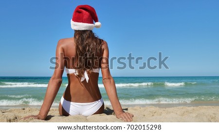 Beautiful girl sits with her back to the sea in a Santa Claus cap, in a white bathing suit, a background of sand and sea blue water

