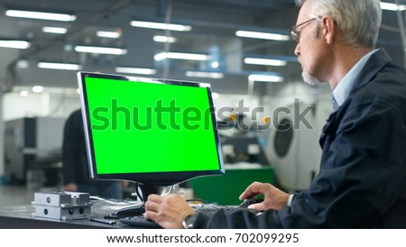Senior engineer in glasses is working on a desktop computer with a green screen on monitor in a factory.