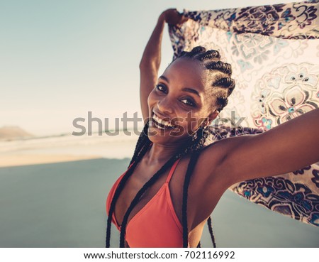 African woman in bikini on the beach holding scarf in the air. Young female with scarf looking at camera.