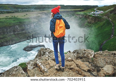 Girl in warm clothing, in red knitted hat and small orange backpack stands on the cliff on background of Gullfoss waterfall in Iceland. Travel to the Iceland