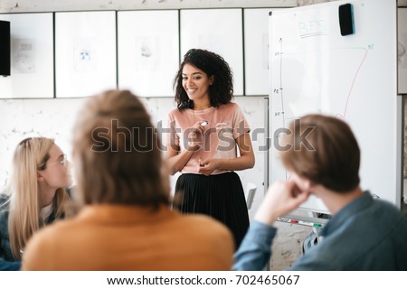 Portrait of cheerful African American lady with dark curly hair standing near board and happily speaking with colleagues in office. Young beautiful business woman giving presentation to coworkers Royalty-Free Stock Photo #702465067