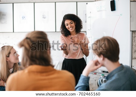 Beautiful African American lady with dark curly hair happily speaking about new project with her colleagues in office. Young smiling business woman giving presentation to coworkers Royalty-Free Stock Photo #702465160