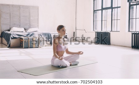 A beautiful, smart, athletic girl does yoga exercises in a gym in loft style, with natural light from large windows. She is dressed in a sporty uniform, breeches and crocheted top. Stretching