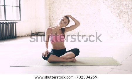 A beautiful, smart, athletic girl does yoga exercises in a gym in loft style, with natural light from large windows. She is dressed in a sporty uniform, breeches and crocheted top. Stretching