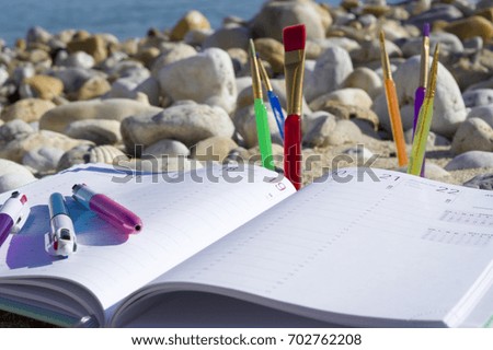 Diary, colorful pens, watercolor paintbrushes on the seashore against
sea stones and the sea
