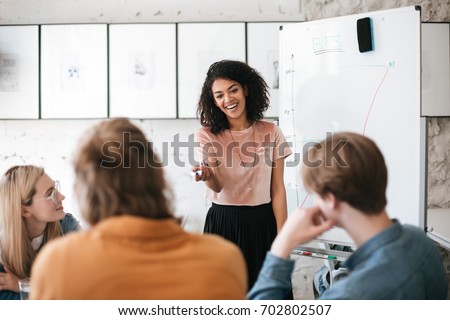 Beautiful African American lady with dark curly hair standing near board and happily discussing new project with her colleagues in office. Young smiling business woman giving presentation to coworkers