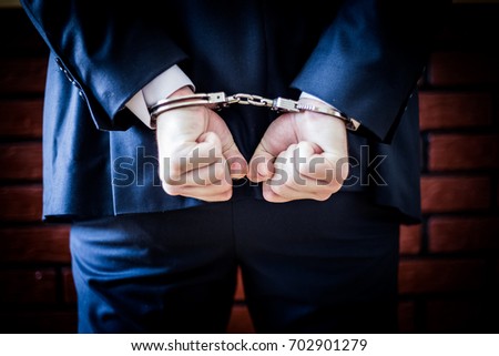 Businessman with fist string hands on the back and handcuffs on. Arrested for corruption and crime in business world. Professor with cuffs corrupted with bricks in the background