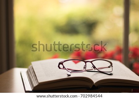 Background for greeting on Teacher's Day. Glasses lie on an open book on a desk. In a background there is a blurred view from a window: green nature and red flowers. 