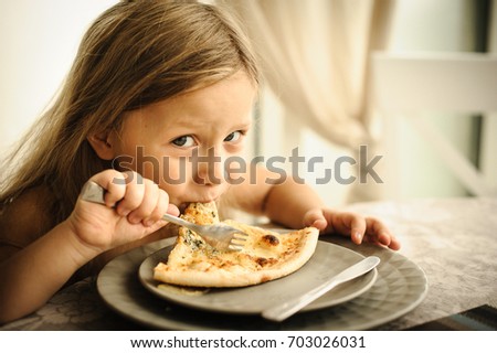 Cute happy young girl concentrated on eating big slice of fresh made pizza. She enjoy sunny day and yummy meal. She has long blonde hair. Close up.