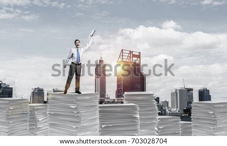 Businessman keeping hand with book up while standing on pile of paper documents with cityscape and sunlight on background. Mixed media.