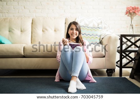 Fun woman in her 20s sitting on the floor of the living room of her apartment eating a bowl of cereal