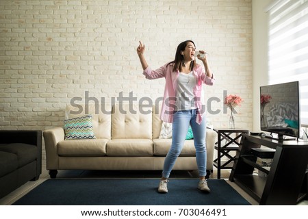 Independent woman singing karaoke alone in her apartment and having fun in the living room Royalty-Free Stock Photo #703046491