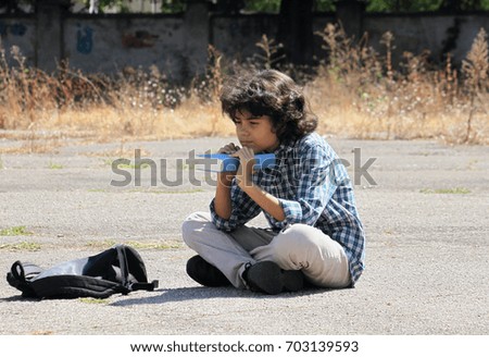 A handsome schoolboy with curly hair is sitting on the ground with a book in his hands