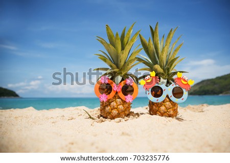 Couple of funny attractive pineapples in children's cheerful sunglasses on sand against turquoise sea. Tropical summer vacation concept. Happy sunny day on the beach of tropical island. 
