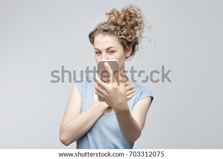 Selective focus on phone. Lady with curly hair in bun in grey t-shirt making selfie using modern mobile phone,looking flirty at camera, posing against white studio wall background. Technology concept.
