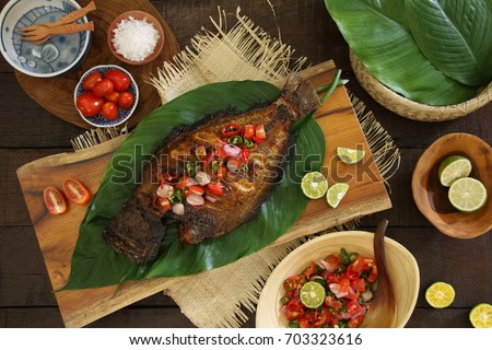 Ikan Bakar Dabu-Dabu. Popular dish of grilled fish with fresh and spicy tomato salsa from Manado. Served on a wooden block lined with broad leaves. Surrounded with extra salsa and other condiments. Royalty-Free Stock Photo #703323616