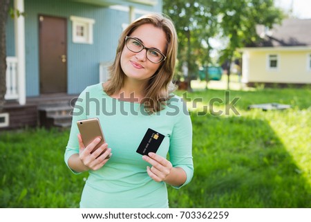 Woman of 30 years old near the house holds a phone and a credit card. Idea for app for renting or buying a house, an apartment.