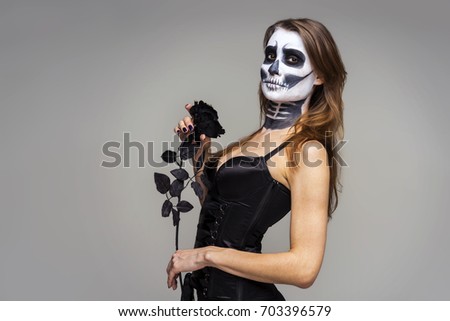 Portrait of woman with halloween skeleton makeup holding black rose flower over gray  background. 