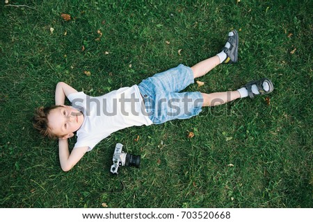 handsome caucasian little boy wearing T-shirt and shorts lying on the green grass with fallen leaves near film camera