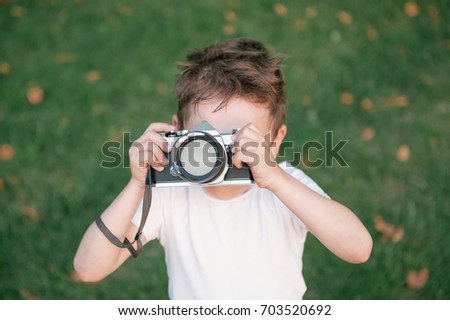 little boy taking a picture using vintage film camera on green grass and fallen leaves background in autumn