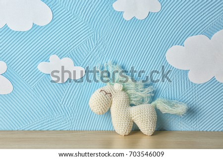 Cute knitted toy horse on wooden table near blue wall with paper clouds