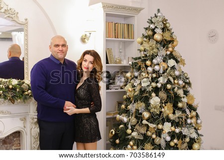 Beautiful spouses, loving husband and wife smile and look at camera, pose and hug each other in bright living room with fireplace decorated in festive. Woman of European appearance with long curly