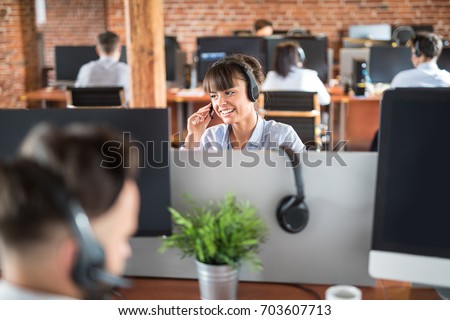 Call center worker accompanied by her team. Smiling customer support operator at work. Young employee working with a headset. Royalty-Free Stock Photo #703607713