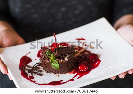 Customer holds chocolate cake with cherry jam, decorated by sweet red berry, almond and mint. Delicious dessert serving in restaurant, close up picture